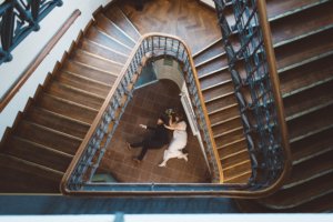 Hochzeitsfoto in Rostock aus der Vogelperspektive: Ein Brautpaar liegt entspannt im Zentrum eines kunstvoll geschwungenen, hölzernen Treppenhauses mit charmantem Altbauflair.