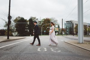 Ein Brautpaar überquert lachend eine leere Straße in Rostock – die Braut in einem fließenden Kleid mit Blumenkranz, der Bräutigam stilvoll im Anzug und Hut, umgeben von städtischer Kulisse und Straßenbahnschienen.