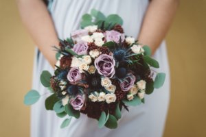 Detailaufnahme eines Hochzeitsstraußes mit cremefarbenen und fliederfarbenen Rosen, blauen Disteln und Eukalyptusblättern, gehalten von einer Braut in fließendem Kleid – aufgenommen in Rostock.