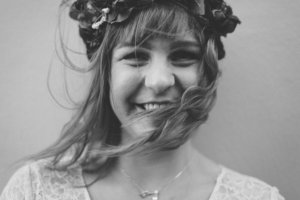 Schwarz-weißes Nahporträt einer lachenden Braut mit Blumenkranz, vom Wind zerzaustes Haar im Gesicht – aufgenommen bei einer Hochzeit in Rostock.