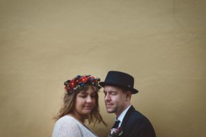 Ein Brautpaar steht eng aneinander gelehnt vor einer warmen, ockerfarbenen Wand in Rostock – beide mit geschlossenen Augen, sie mit Blumenkranz, er mit schwarzem Hut und Anstecker.