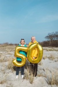 Paarshooting zur Goldenen Hochzeit am Strand in Markgrafenheide Ein älteres Paar steht auf einer sandigen Dünenlandschaft in Rostock und hält große goldene Ballons in Form der Zahl 50 – als Symbol ihrer goldenen Hochzeit unter blauem Himmel.