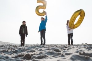 Strandfotos mit Kindern in Rostock – Generationen feiern die goldene Hochzeit Drei Kinder stehen am Strand von Rostock mit goldenen Zahlenballons in Form der Zahl 50 – ein verspielter Moment zum Jubiläum der Großeltern bei sonnigem Wetter und maritimer Kulisse.