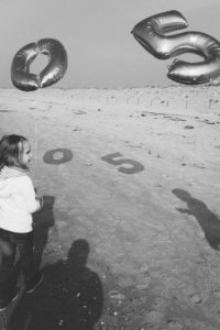 Familienshooting am Strand – Schattenspiel mit Jubiläumsballons zur goldenen Hochzeit Schwarz-Weiß-Aufnahme eines Familienshootings am Strand: Ein Kind hält einen goldenen Ballon in Form der Zahl 0, daneben schwebt die Zahl 5 – deren Schatten projizieren die Zahl 50 auf den Sand.
