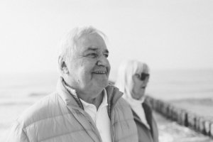 Familienshooting am Strand – Persönliches Porträt zum 50. Ehejubiläum Schwarz-Weiß-Porträt eines älteren Mannes, der am Strand von Rostock lächelt – im Hintergrund seine Frau, beide Teil eines Familienshootings zur Feier ihrer goldenen Hochzeit.