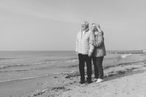 Goldene Hochzeit am Strand von Rostock mit natürlichen Paarfotos Ein älteres Ehepaar steht eng umschlungen am Strand von Rostock, schaut lächelnd aufs Meer – aufgenommen als Paarshooting zur goldenen Hochzeit in stilvollem Schwarz-Weiß.