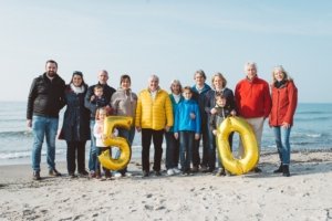 Familienshooting am Strand mit Mehrgenerationen-Familienfoto zur goldenen Hochzeit Familienshooting am Strand mit einer große Familie mit mehreren Generationen. Stehen bei sonnigem Wetter am Strand, halten goldene Ballons in Form der Zahl 50 und feiern goldene Hochzeit gemeinsam.