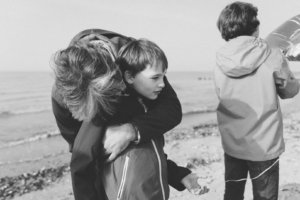 Inniger Moment zwischen Vater und Sohn am Strand Ein Vater umarmt liebevoll seinen Sohn am Strand. Der Junge blickt gedankenversunken in die Ferne, während ein weiteres Kind im Hintergrund mit einem Ballon spielt. Die Szene ist in Schwarz-Weiß aufgenommen und strahlt emotionale Nähe aus.