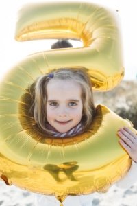 Mädchen schaut durch goldenen Ballon in Form einer Fünf am Strand Ein blondes Mädchen blickt fröhlich durch die Mitte eines großen, goldenen Luftballons in Form der Zahl 5. Im Hintergrund scheint die Sonne, der Strand ist leicht zu erkennen. Die Szene strahlt Unbeschwertheit und kindliche Freude aus.