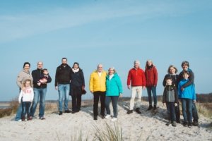 Großfamilie auf einer Düne in Markgrafenheide beim Familienshooting in Rostock während des gemeinsamen Strandurlaub Eine Großfamilie mit mehreren Generationen steht bei sonnigem Wetter auf einer Düne am Strand. Alle tragen farbenfrohe Outdoor-Kleidung und genießen sichtlich die gemeinsame Zeit in der Natur. Im Hintergrund ist blauer Himmel und Küstenlandschaft zu sehen.