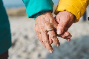 Händchen haltendes älteres Paar am Strand – Zeichen lebenslanger Liebe zur goldenen Hochzeit Nahaufnahme zweier Hände, die sich liebevoll halten. Eine Hand trägt einen Ehering, beide tragen farbige Jacken (grün und gelb). Der Hintergrund zeigt unscharf den Sandstrand – ein Symbol für Zusammenhalt und Verbundenheit im Alter.